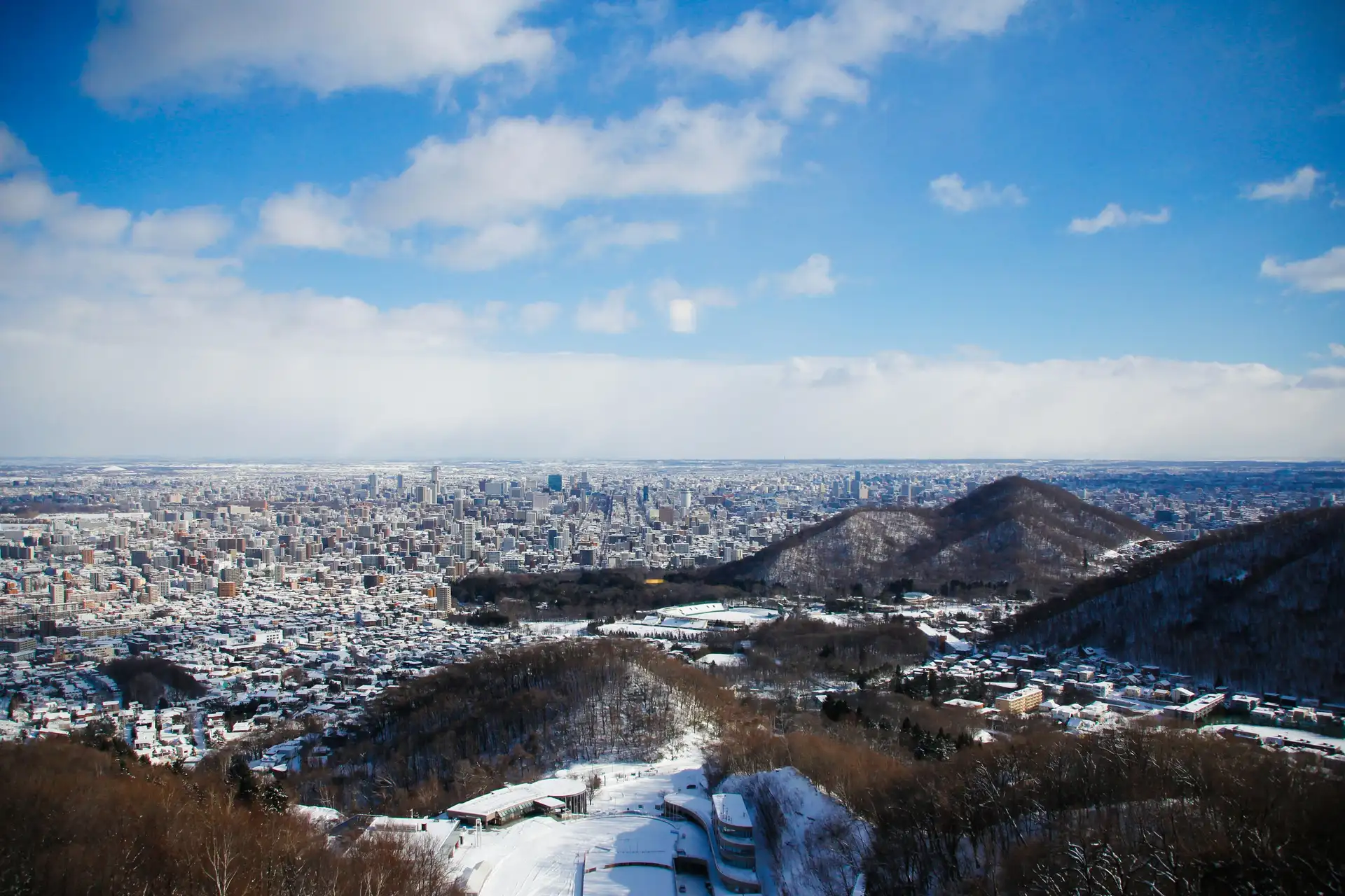 Vue panoramique en plongée sur la ville de Sapporo enneigée depuis une colline, sous un ciel bleu parsemé de nuages blancs à Hokkaido.