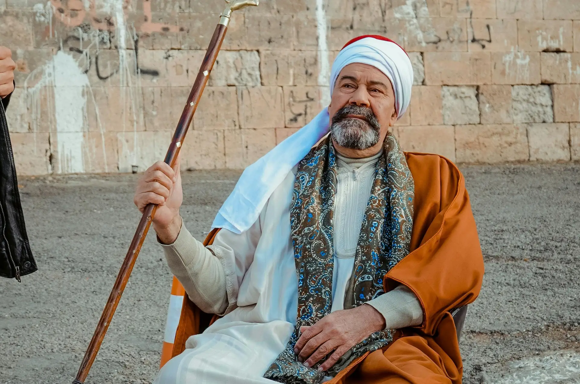 Voyage en Egypte - Homme en tenue traditionnelle assis avec une canne devant un mur en pierre