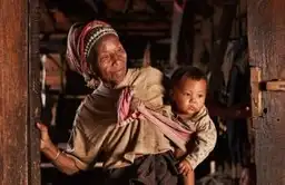 Voyage au Laos — Une femme locale portant un enfant sur son dos devant l'entrée d'une maison traditionnelle en bois.