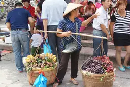 Une marchande de rue portant deux paniers de fruits à l'aide d'un fléau en bois sur son épaule