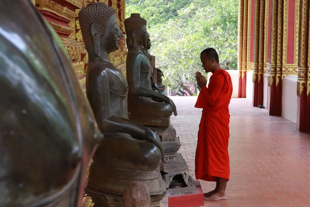 Moine bouddhiste en robe orange se recueillant mains jointes devant un alignement de statues de Bouddha dans un temple au Laos.