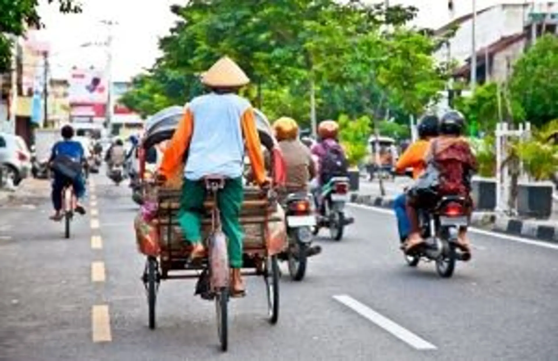 Conducteur de becak traditionnel et scooters circulant sur une avenue arborée dans une ville d'Indonésie.