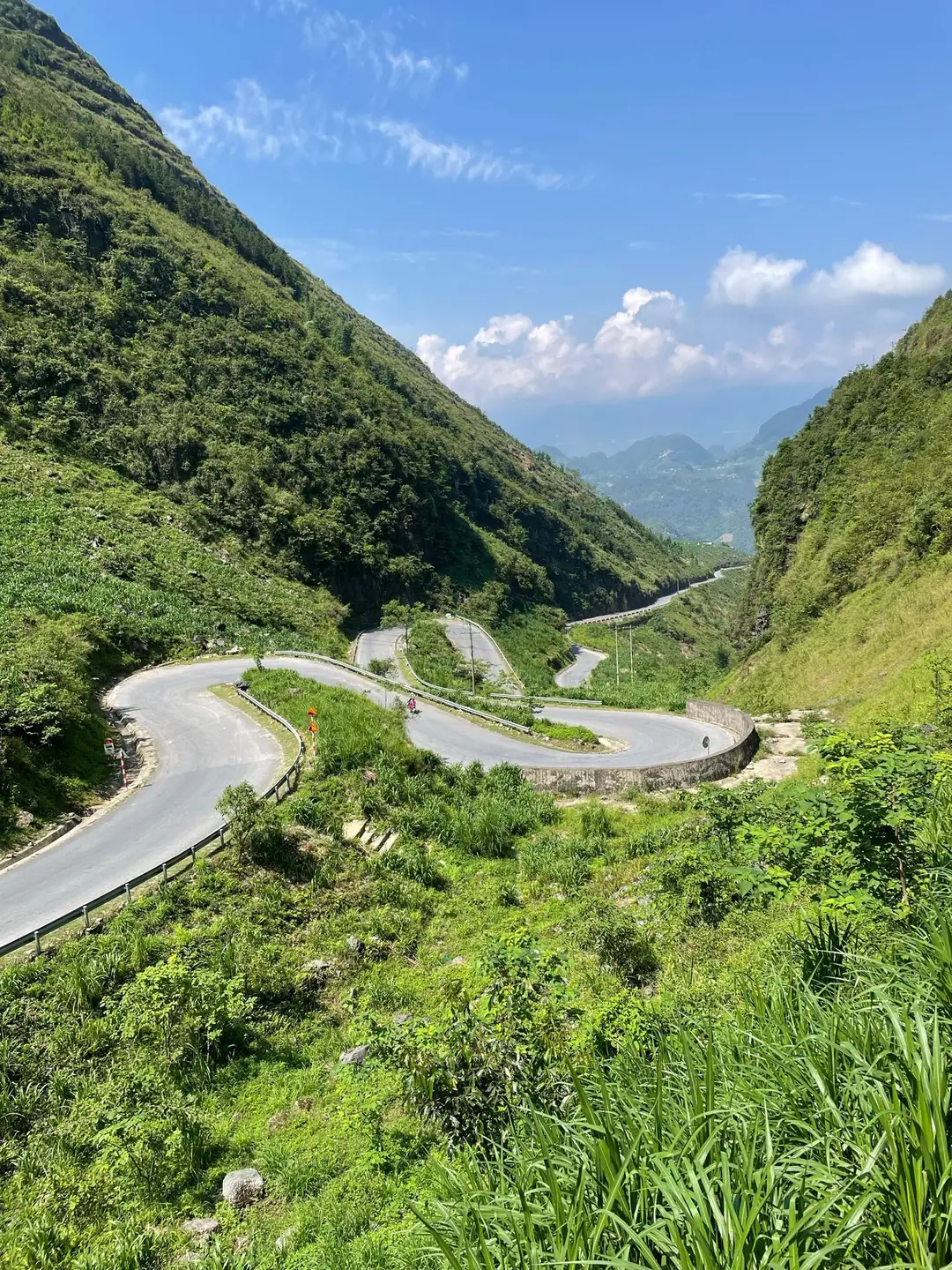Une route asphaltée en lacets serrés serpente à flanc de montagne verdoyante sous un ciel bleu parsemé de nuages dans la province d'Ha Giang.