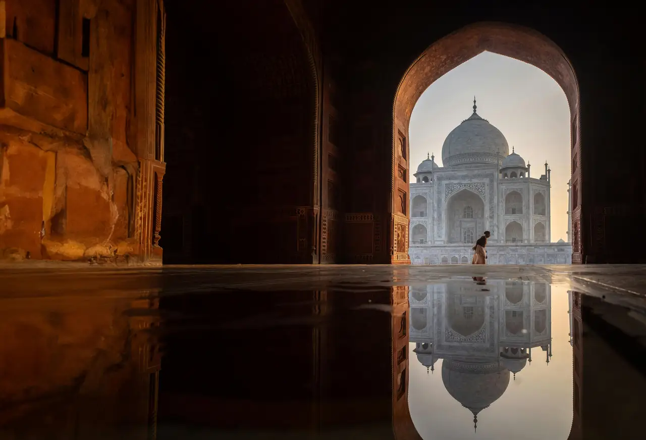 Silhouette du Taj Mahal vue à travers une arche en pierre au coucher du soleil avec un reflet parfait dans une étendue d'eau calme à Agra.