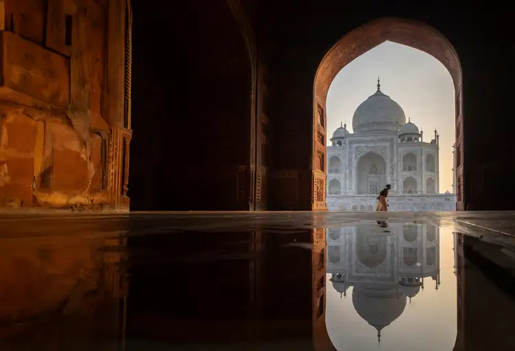 Silhouette du Taj Mahal vue à travers une arche en pierre au coucher du soleil avec un reflet parfait dans une étendue d'eau calme à Agra.