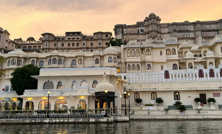 Façade illuminée du City Palace d'Udaipur surplombant les eaux calmes du lac Pichola sous un ciel de coucher du soleil aux teintes orangées.