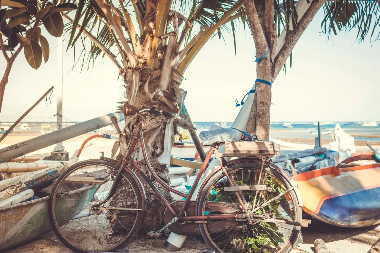 Une bicyclette traditionnelle appuyée contre un tronc de cocotier sur une plage bordée de pirogues locales à Bali.