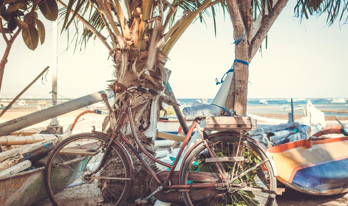 Voyage en Asie — Une vieille bicyclette rouillée appuyée contre un palmier sur une plage balinaise, avec un bateau traditionnel et la mer en arrière-plan, ambiance authentique et paisible.