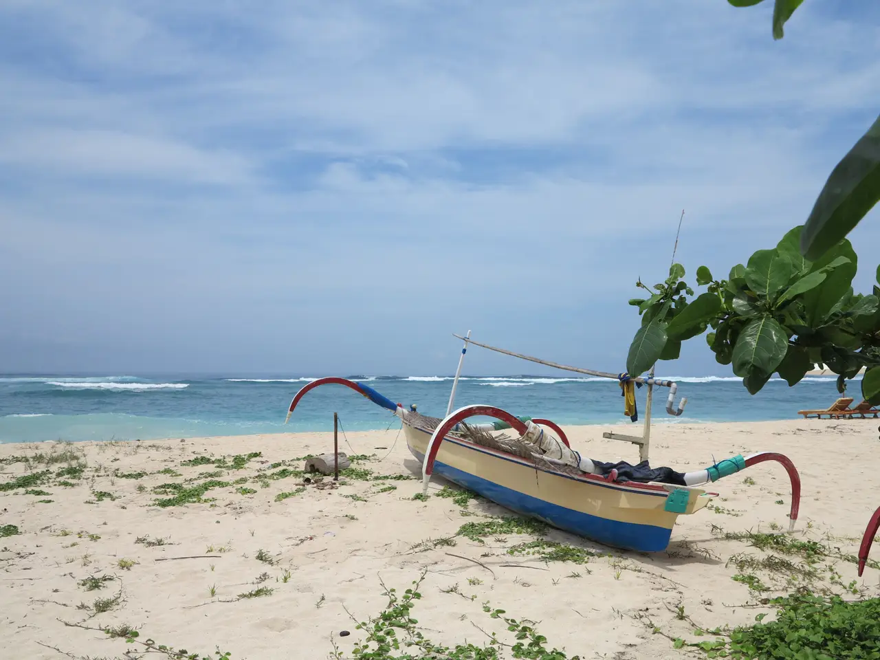 Une pirogue traditionnelle balinaise posée sur le sable d'une plage ombragée par de la végétation tropicale, face à une mer calme sous un ciel légèrement nuageux à Candidasa.