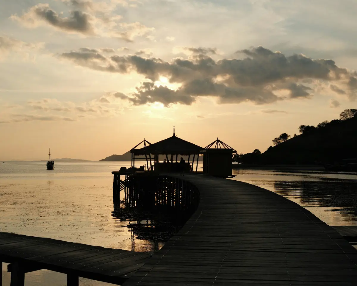 Ponton en bois s'avançant sur une mer calme à Nusa Lembongan au coucher du soleil, avec la silhouette d'une cabane de plage sous un ciel nuageux.