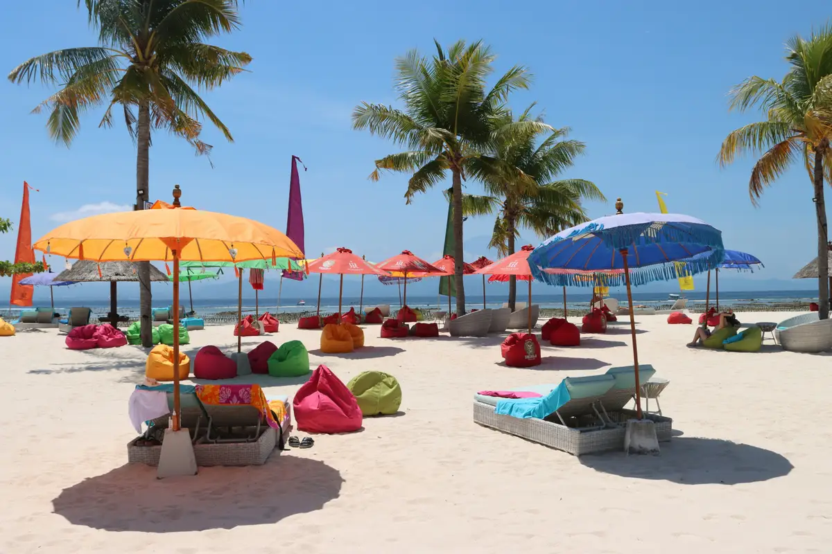 Une plage de sable blanc à Nusa Lembongan parsemée de parasols colorés et de poufs de plage, bordée de grands palmiers sous un ciel bleu azur.