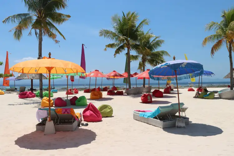 Une plage de sable blanc à Nusa Lembongan parsemée de parasols colorés et de poufs de plage, bordée de grands palmiers sous un ciel bleu azur.