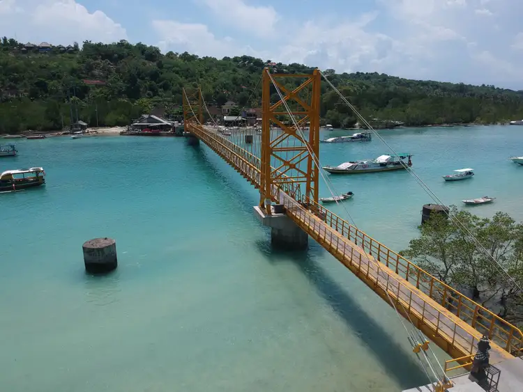 Vue aérienne du célèbre pont jaune suspendu reliant Nusa Lembongan et Nusa Ceningan, enjambant une eau turquoise parsemée de petits bateaux.