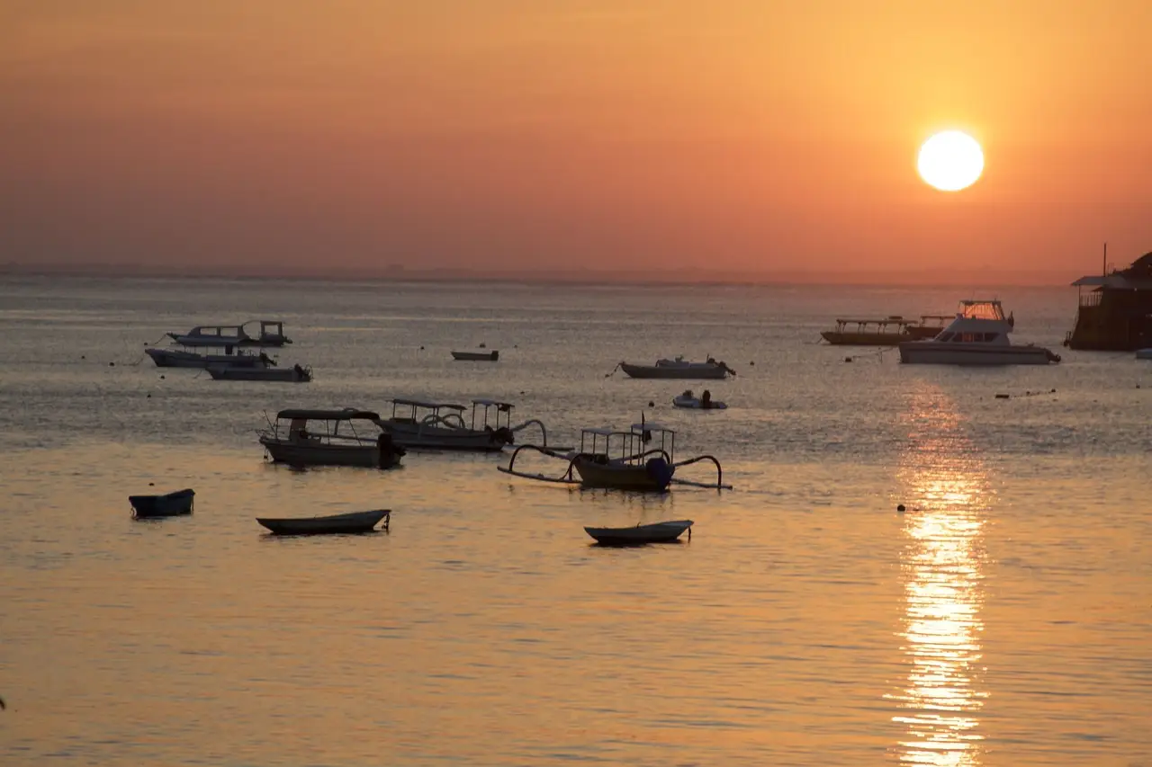 Plusieurs bateaux traditionnels à balanciers ancrés près de la côte de Jimbaran face à un grand soleil couchant embrasant le ciel de teintes orangées à Bali.