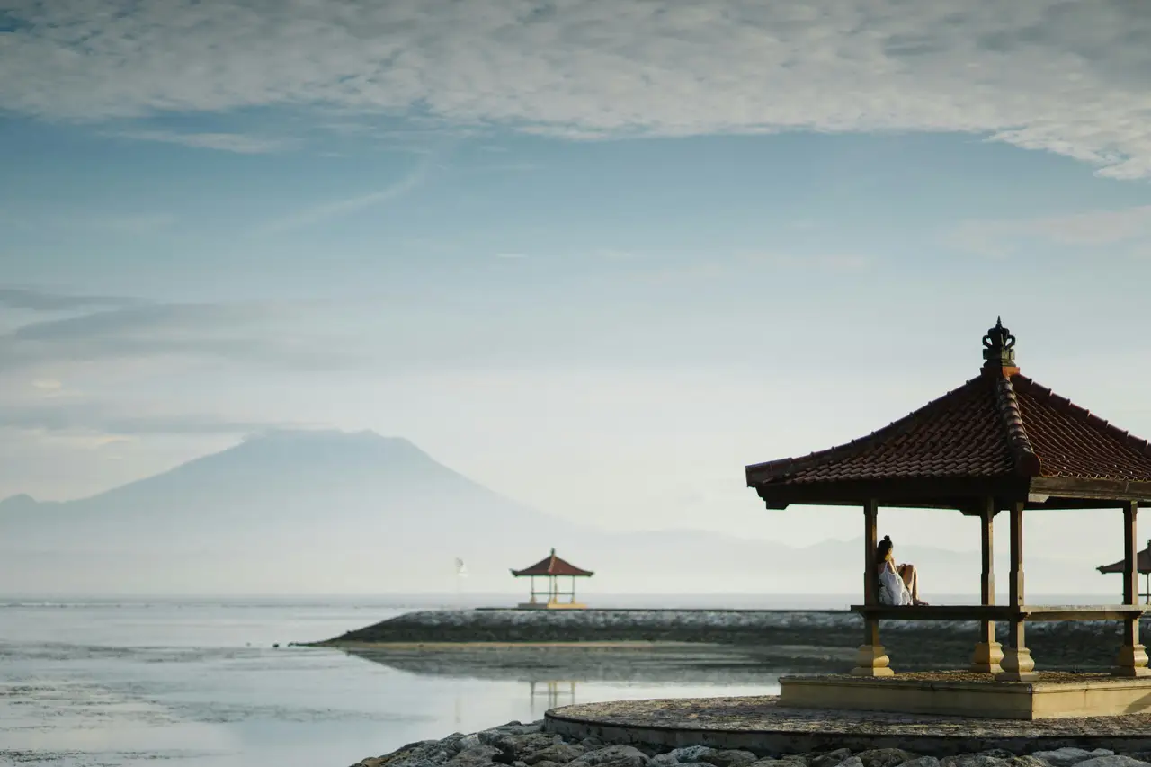 Un pavillon balinais traditionnel sur pilotis au bord de l'eau à Sanur, offrant une vue sur la mer calme avec une montagne majestueuse se dessinant à l'horizon sous un ciel voilé.