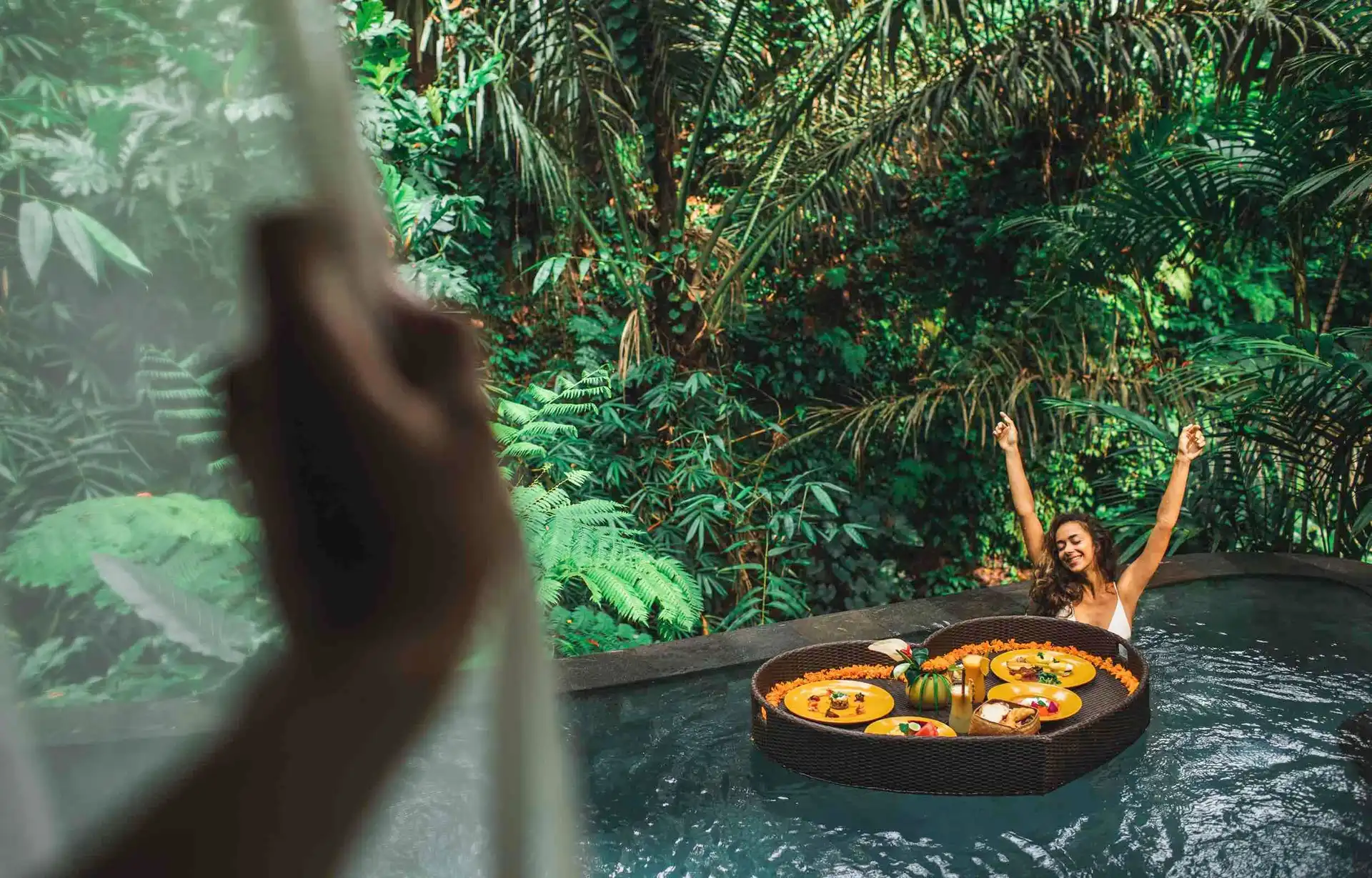 Travel in Asia - A woman with arms raised in happiness in a pool about to enjoy a floating breakfast served on a heart-shaped tray at a resort in Bali
