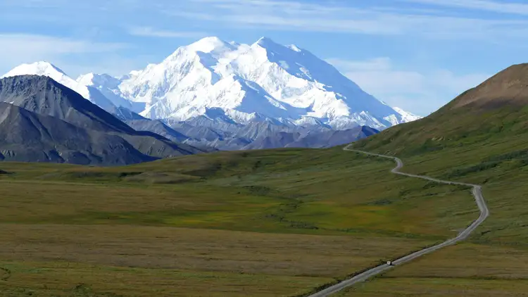 Route sinueuse traversant la toundra verdoyante menant vers le sommet enneigé du mont Denali sous un ciel bleu dégagé