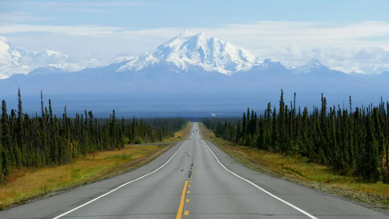 Route rectiligne traversant une forêt boréale de conifères vers un imposant volcan ou sommet enneigé sous un ciel clair en Alaska
