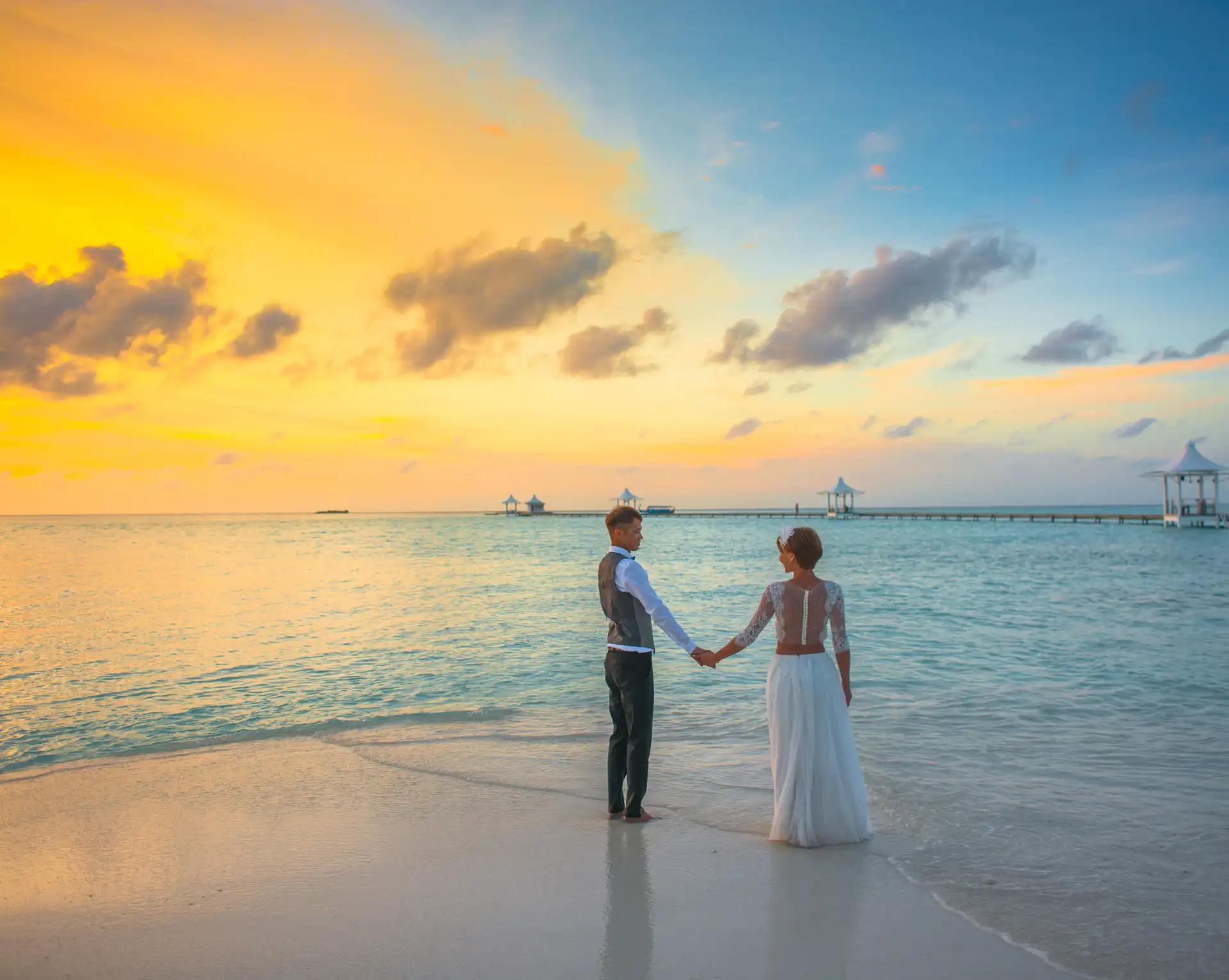 Couple de mariés se tenant par la main au bord de la mer sur une plage tropicale au coucher du soleil.