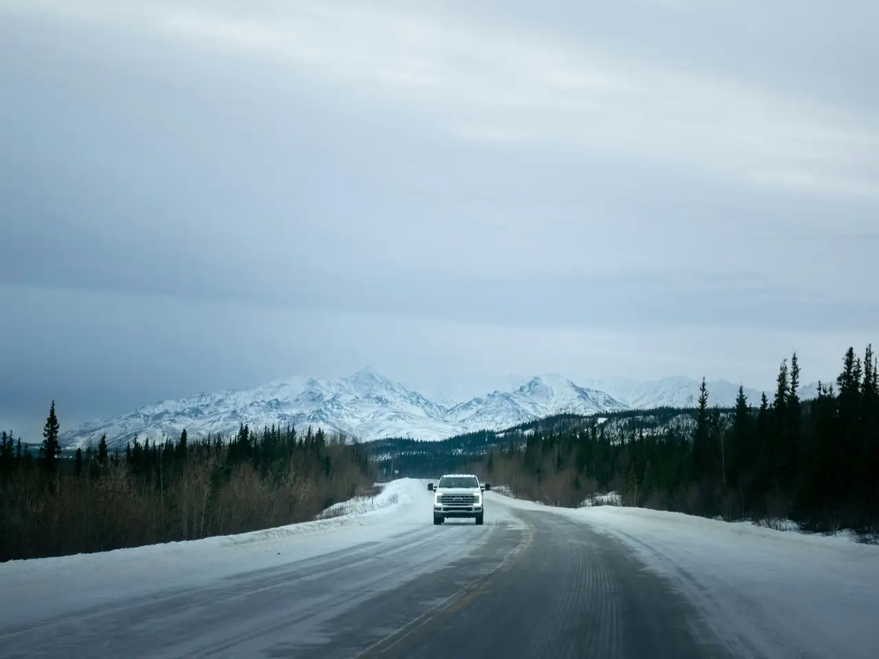 Route enneigée traversant une forêt de conifères face à une chaîne de montagnes lointaines sous un ciel d'hiver voilé entre Denali et Fairbanks