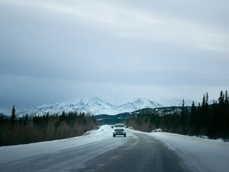 Route enneigée traversant une forêt de conifères face à une chaîne de montagnes lointaines sous un ciel d'hiver voilé entre Denali et Fairbanks