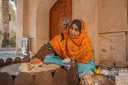 Femme omanaise vêtue d'un voile orange préparant du pain traditionnel sur une plaque de cuisson circulaire.