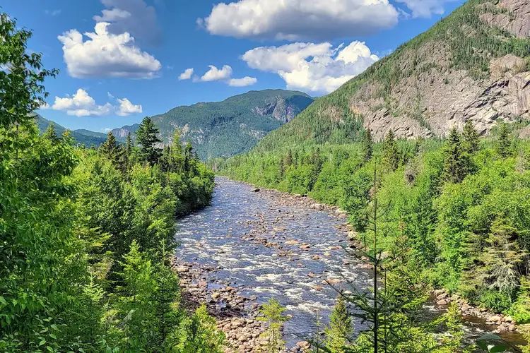 Rivière s'écoulant dans une vallée encaissée bordée d'une forêt dense et de versants montagneux sous un ciel bleu parsemé de nuages dans la région de Charlevoix