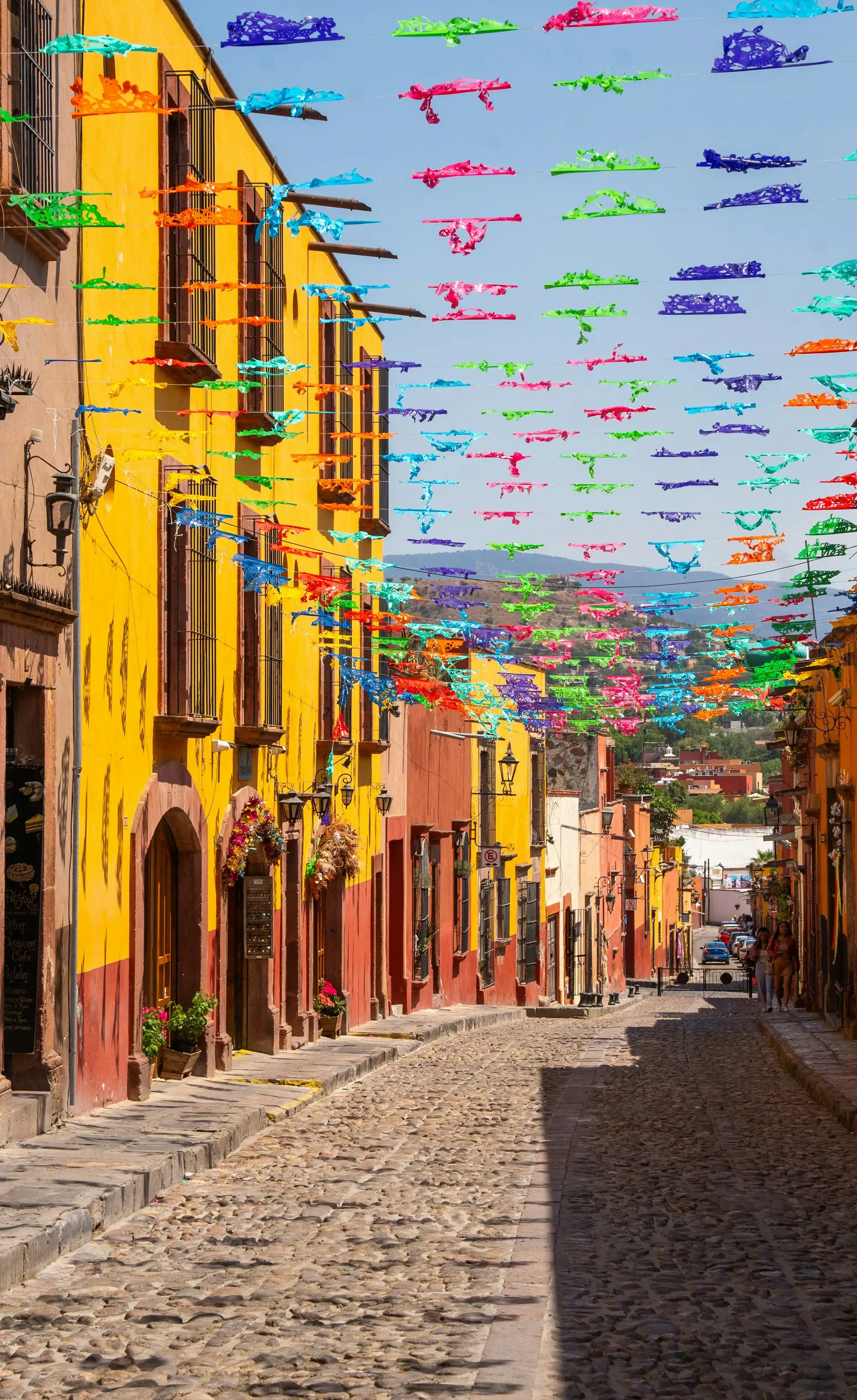Voyage au Mexique – Une ruelle pavée bordée de maisons coloniales aux façades jaunes et orange décorée de guirlandes de fanions colorés sous le soleil.
