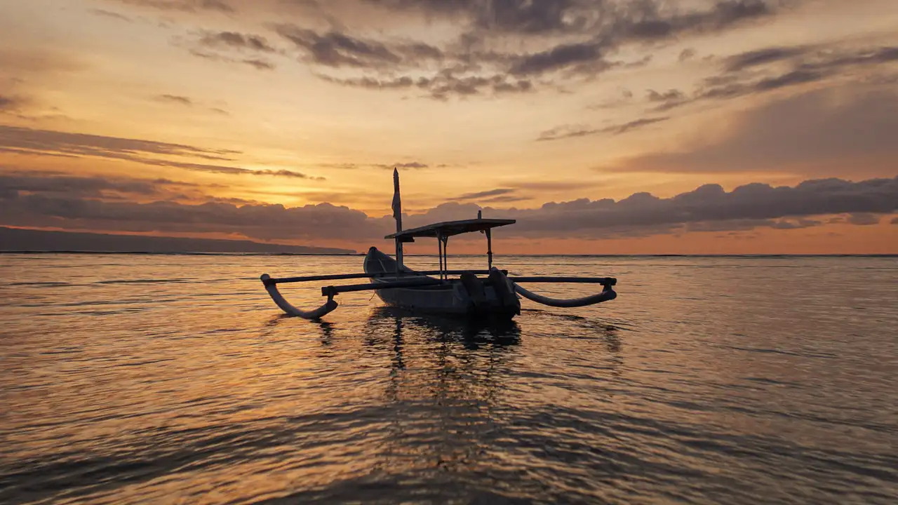 Silhouette d'un jukung, bateau de pêche traditionnel balinais à balanciers, immobile sur une mer calme face à un ciel flamboyant au coucher du soleil à Bali.