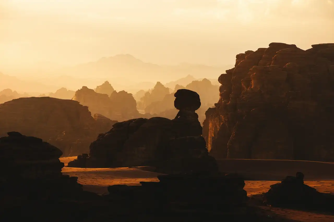 Formations rocheuses majestueuses et falaises désertiques d'AlUla silhouettées contre une lumière dorée et brumeuse au coucher du soleil en Arabie Saoudite.