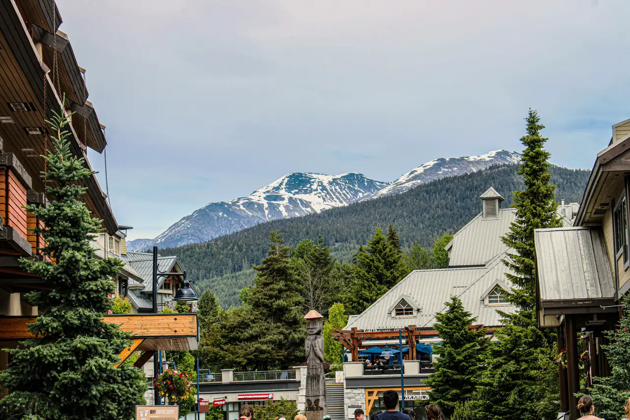 Chalets en bois et sapins du village de Whistler avec en arrière-plan les sommets enneigés de la montagne sous un ciel bleu