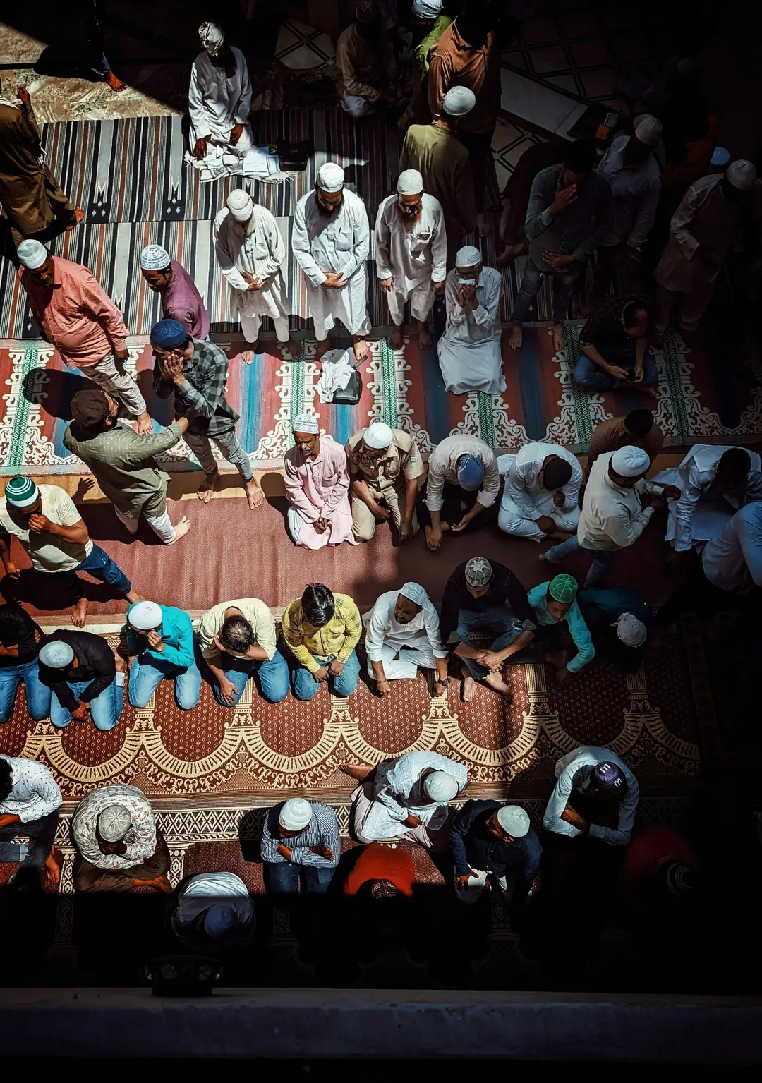 Vue de dessus d'hommes en prière assis sur des tapis colorés dans une mosquée, avec des jeux d'ombres portées et de lumière naturelle intense.