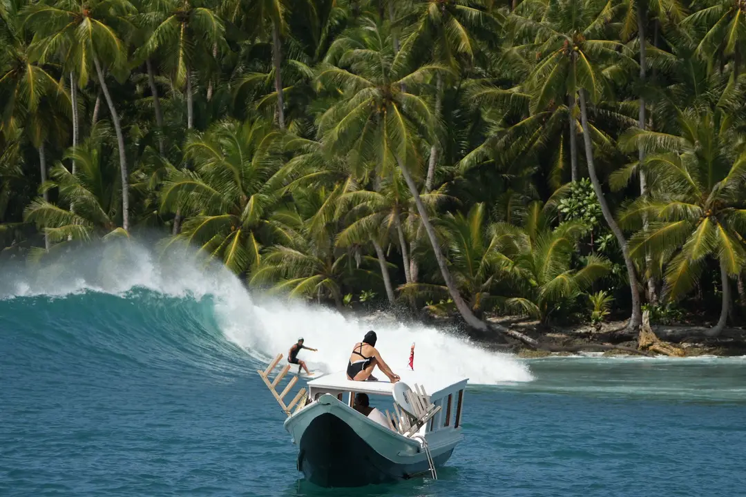 Voyage en Indonésie – Un surfeur glisse sur une vague turquoise aux îles Mentawai à Sumatra, observé depuis un bateau traditionnel devant une côte bordée de cocotiers.