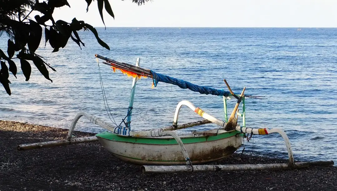 Voyage en Asie – Un jukung, petit bateau traditionnel à balancier, posé sur la plage de sable volcanique noir face à la mer calme à Amed à Bali.