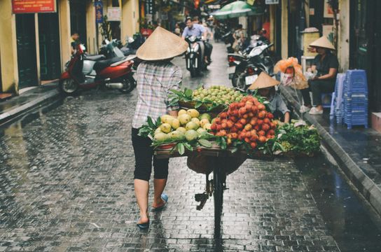 Voyage au Vietnam — Scène de rue à Hanoï avec un vendeur portant un Nón Lá poussant un chariot de fruits et légumes sur une rue pavée humide.