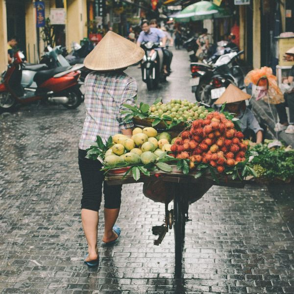 Voyage au Vietnam — Scène de rue à Hanoï avec un vendeur portant un Nón Lá poussant un chariot de fruits et légumes sur une rue pavée humide.