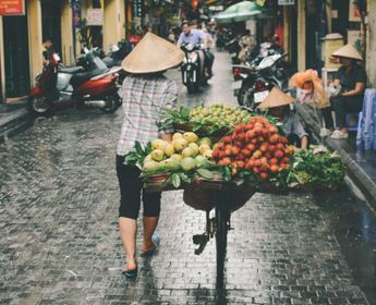 Voyage au Vietnam — Scène de rue à Hanoï avec un vendeur portant un Nón Lá poussant un chariot de fruits et légumes sur une rue pavée humide.