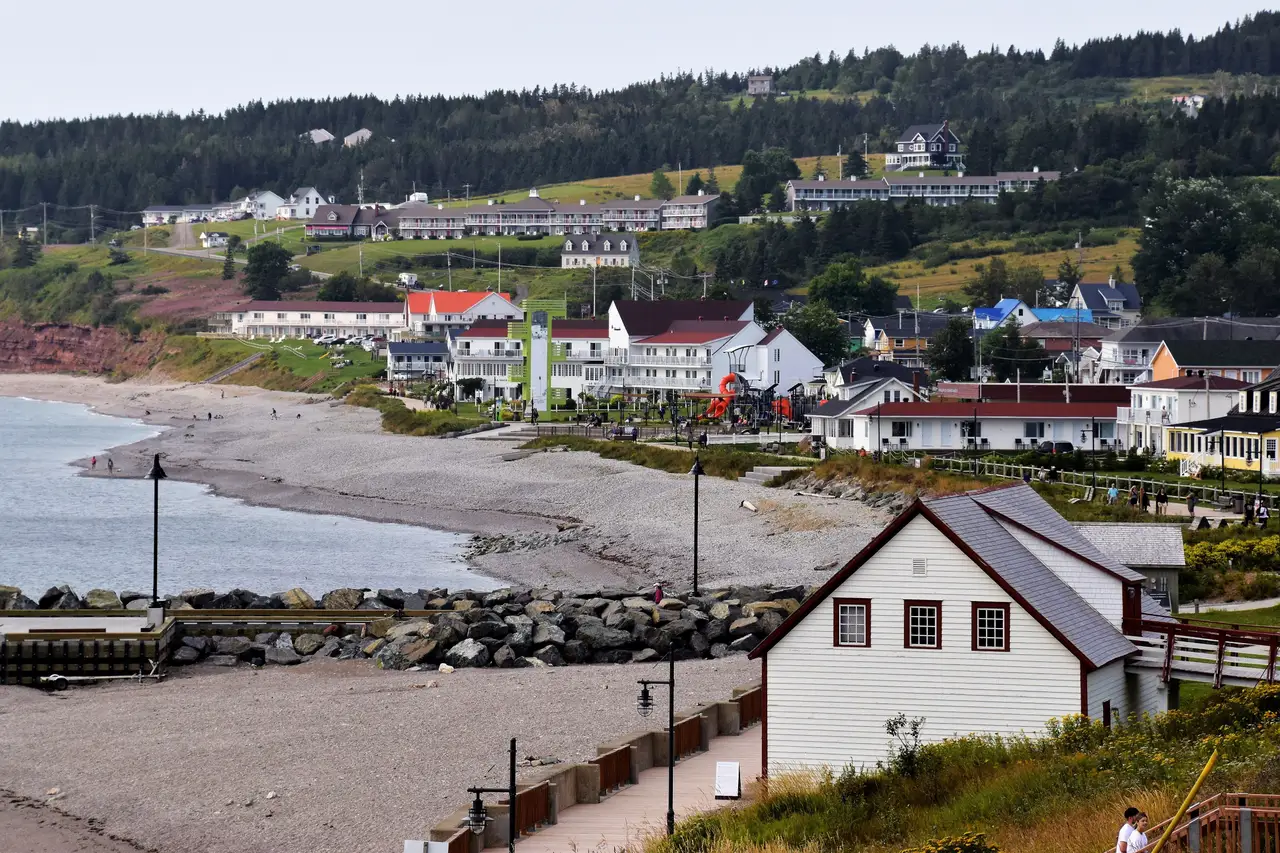Vue plongeante sur les maisons du village côtier bordant l'anse et la plage de galets au pied des collines verdoyantes en Gaspésie