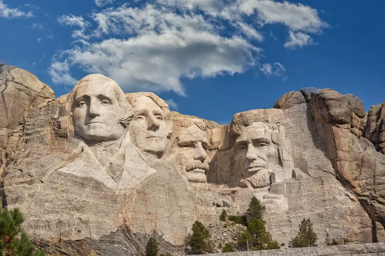 Visages des présidents américains sculptés dans la falaise de granit du mémorial national du Mont Rushmore sous un ciel bleu parsemé de nuages