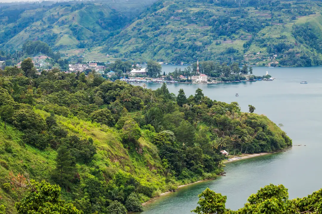 Voyage en Indonésie – Vue en plongée sur le lac Toba, les collines verdoyantes de la caldeira et un village côtier sur les rives de l'île de Samosir à Sumatra.