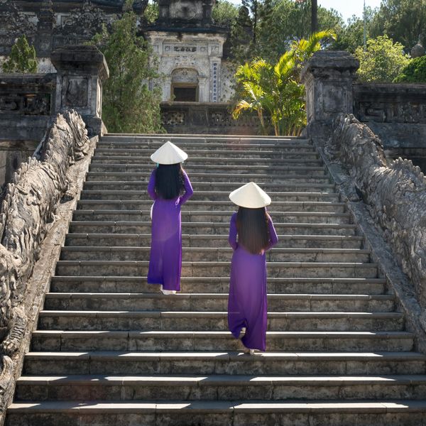 Voyage au Vietnam — Deux femmes en Áo Dài violet et chapeau conique montent l’escalier monumental du Tombeau de Khải Định à Hué, décoré de sculptures de dragons.