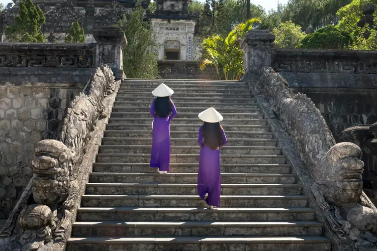 Voyage au Vietnam — Deux femmes en Áo Dài violet et chapeau conique montent l’escalier monumental du Tombeau de Khải Định à Hué, décoré de sculptures de dragons.