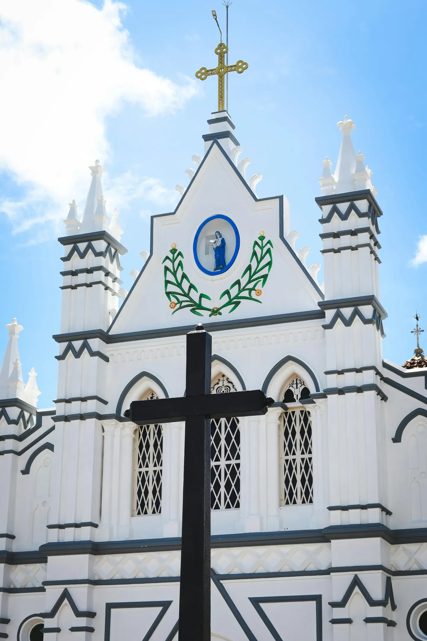 Façade blanche sculptée d'une église chrétienne en Inde, ornée d'une icône de la Vierge Marie et de palmes vertes, avec une grande croix noire au premier plan sur un ciel bleu.