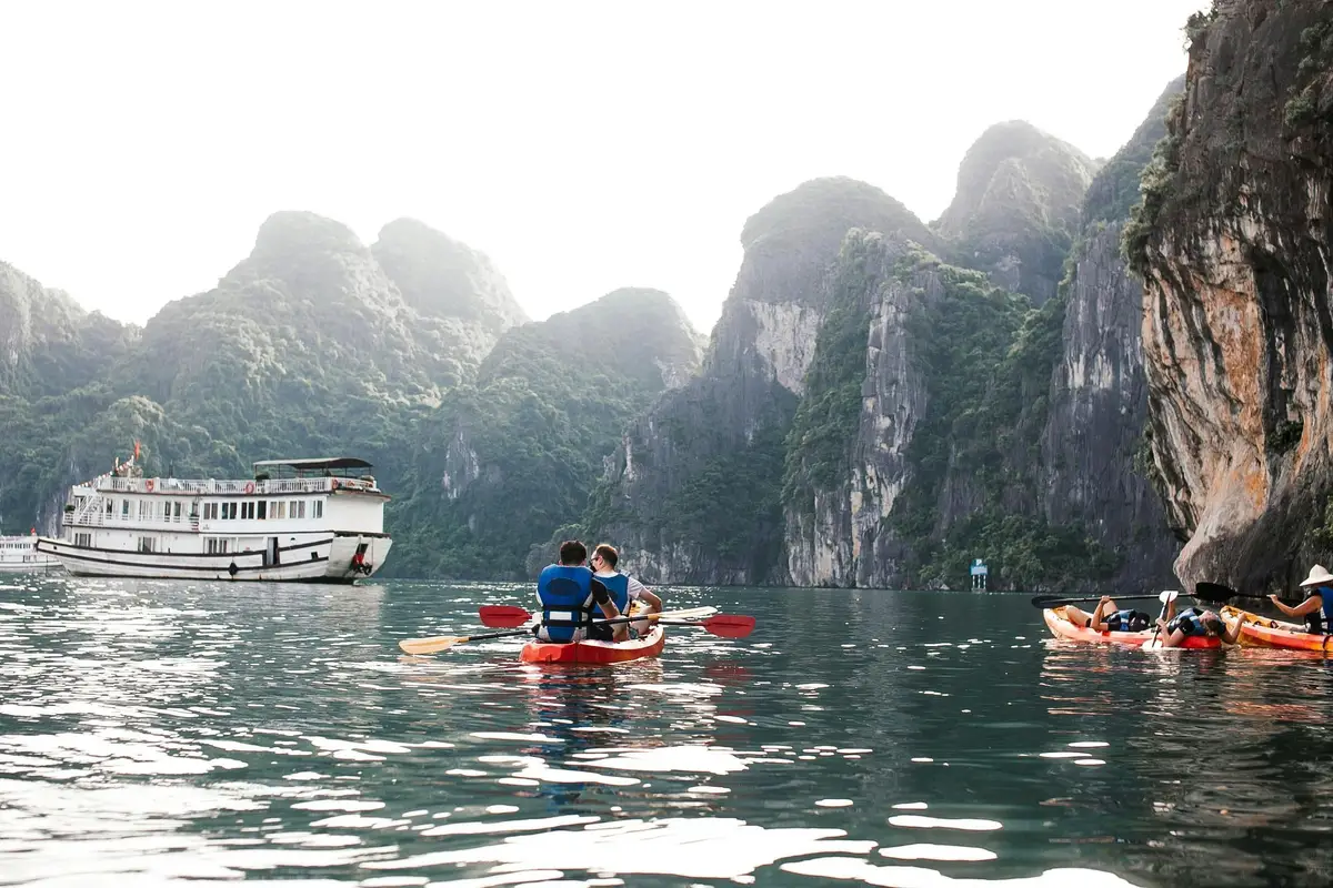 Voyage au Vietnam — Deux personnes en kayak dans la Baie d’Ha Long avec falaises karstiques et bateau de croisière en arrière-plan.