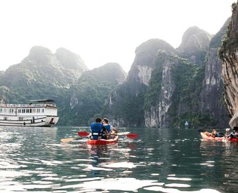 Voyage au Vietnam — Deux personnes en kayak dans la Baie d’Ha Long avec falaises karstiques et bateau de croisière en arrière-plan.