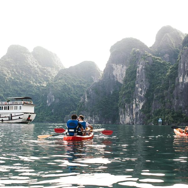 Voyage au Vietnam — Deux personnes en kayak dans la Baie d’Ha Long avec falaises karstiques et bateau de croisière en arrière-plan.