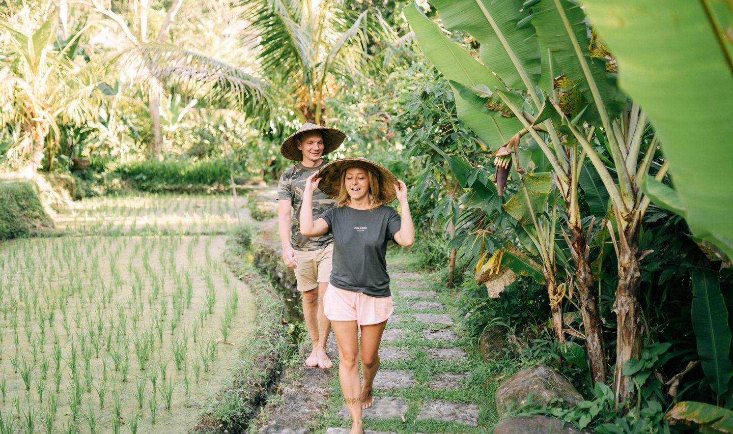 Travel in Asia - A young couple walking on a stone path between a rice field and tropical plants wearing hats