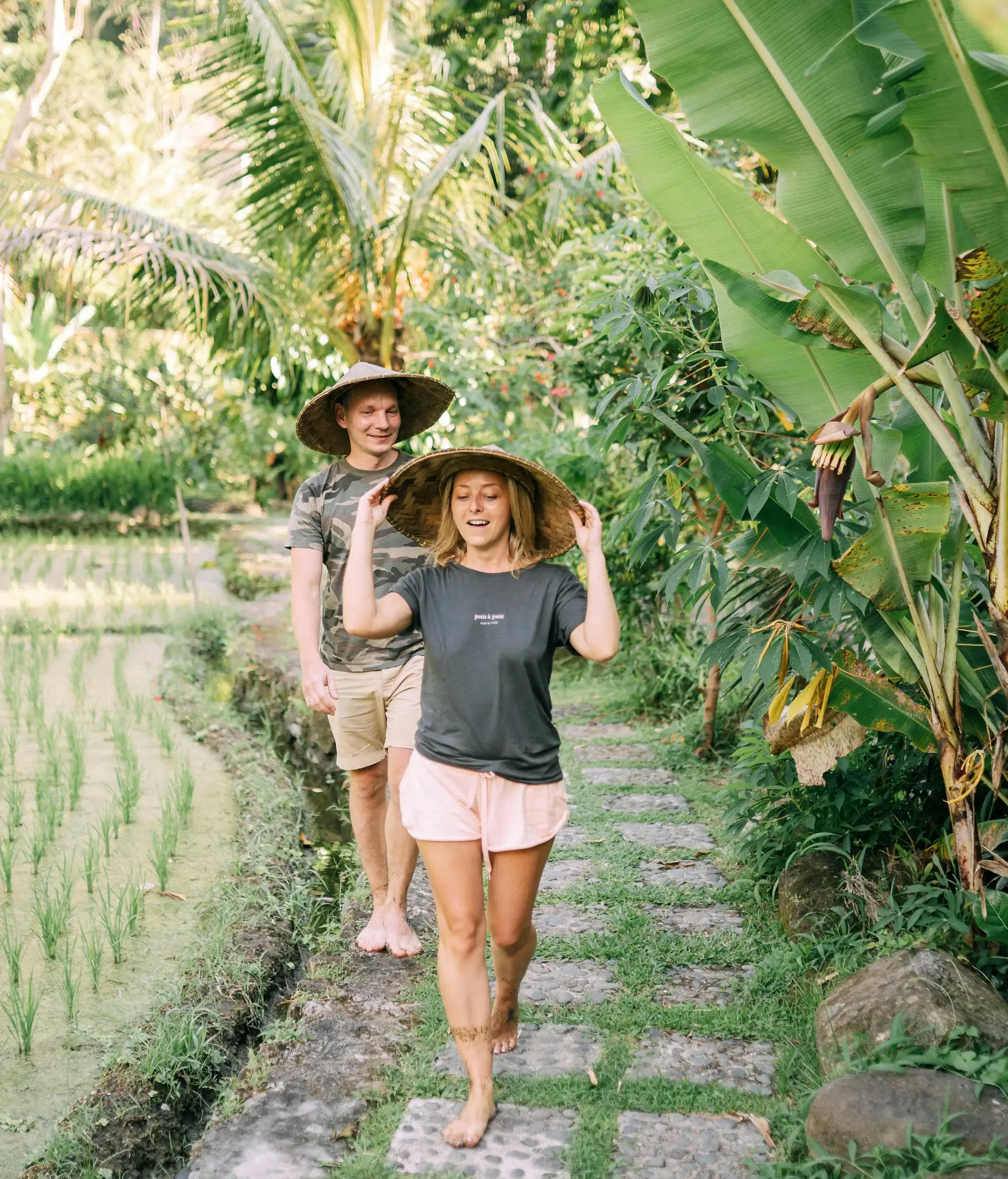 Travel in Asia - A young couple walking on a stone path between a rice field and tropical plants wearing hats