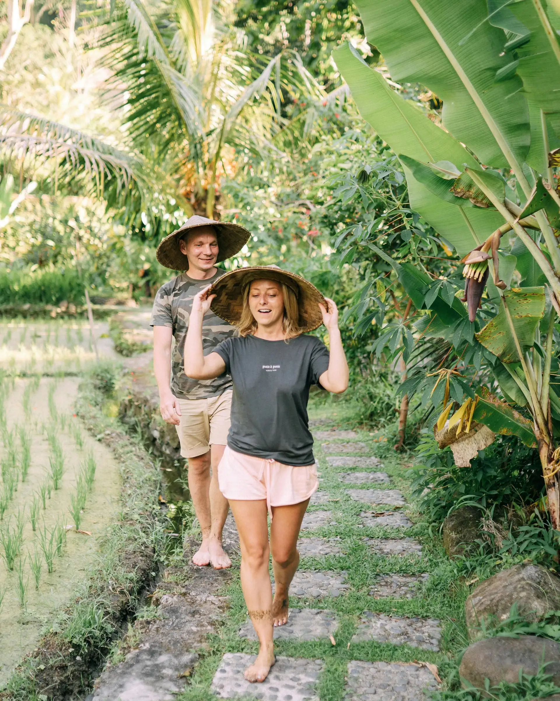 Travel in Asia - A young couple walking on a stone path between a rice field and tropical plants wearing hats