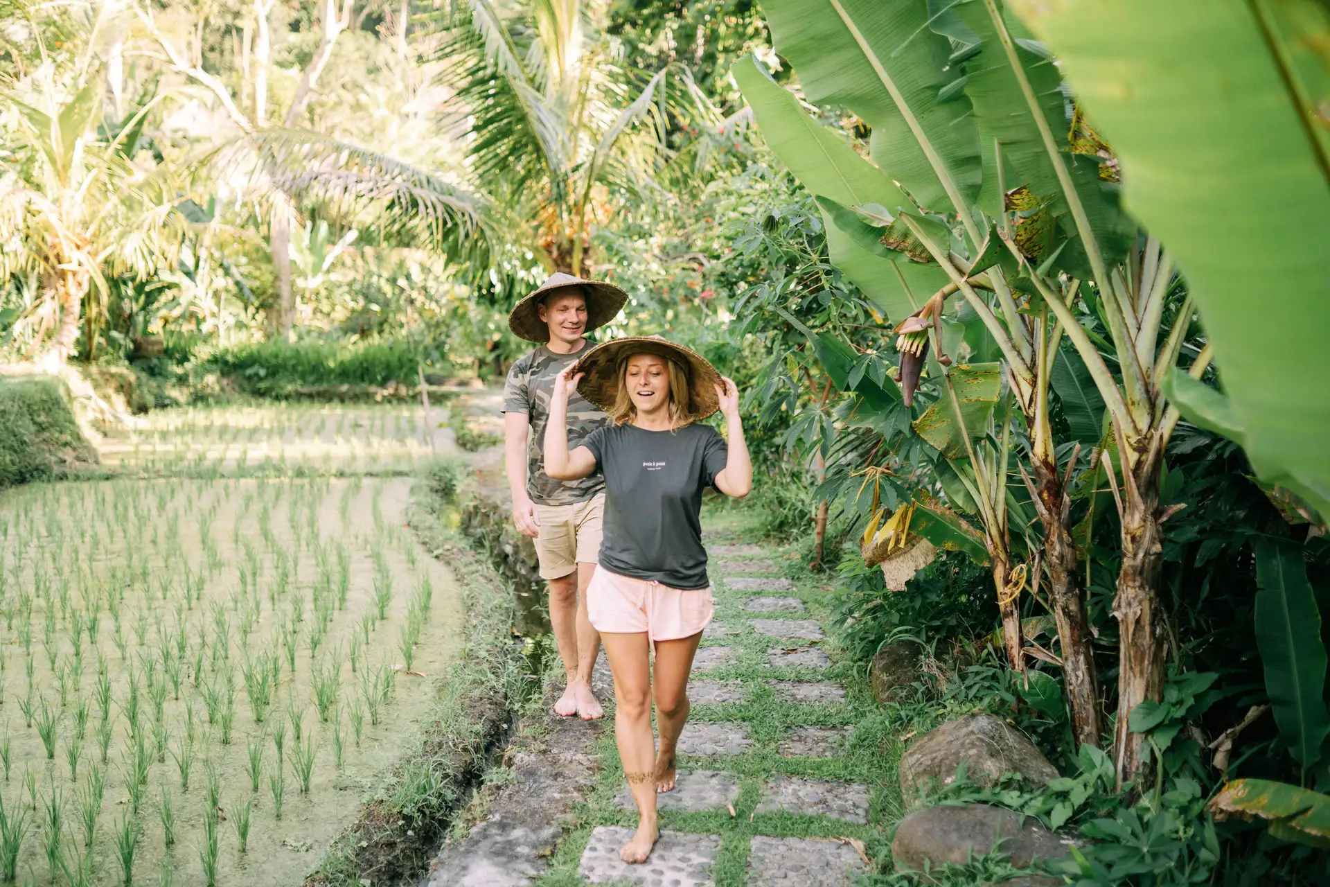 Travel in Asia - A young couple walking on a stone path between a rice field and tropical plants wearing hats